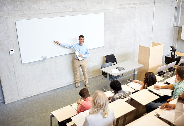 A picture of a classroom with a teacher is pointing at the board and the students are listening.