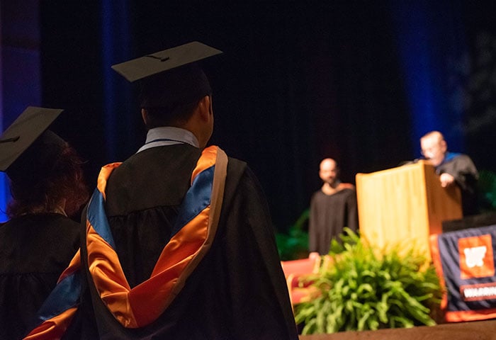 A picture of a graduation event focused on two students listening to their teachers on the stage.
